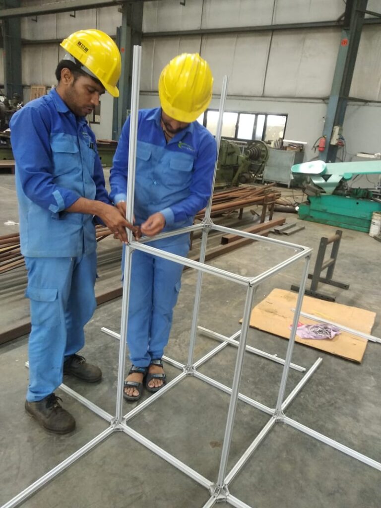 Two workers in blue uniforms and yellow hard hats assembling a rectangular frame out of aluminum extrusions in an industrial workshop.