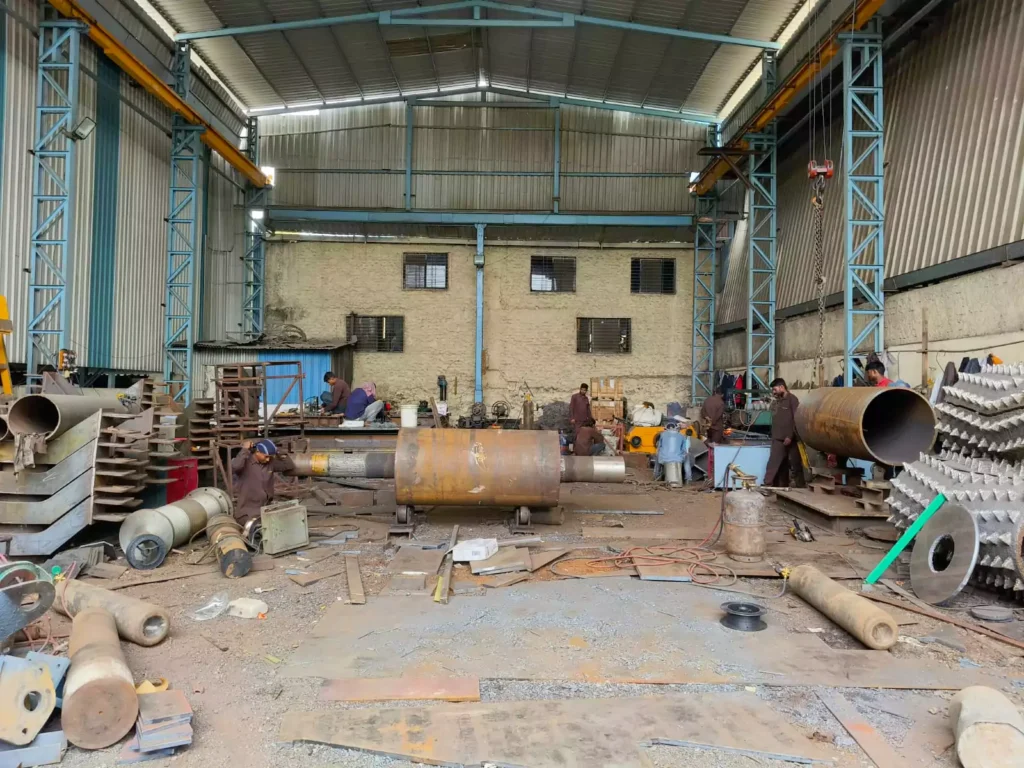 Workers fabricating large heavy metal cylinders and spiked crushing drums in an industrial warehouse workshop with scattered tools and scrap metal.